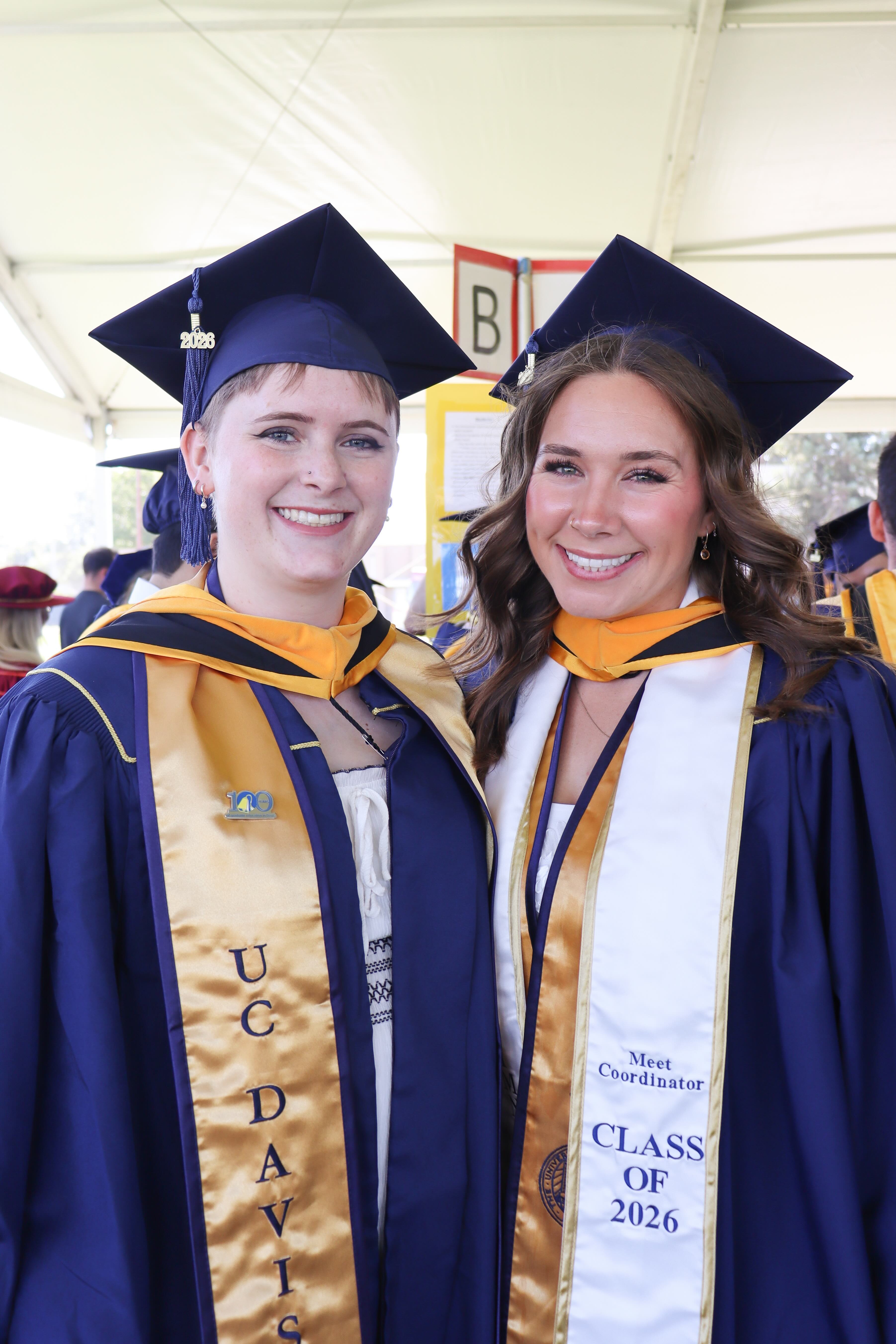 Two smiling Master's graduates in navy caps and gowns with gold stoles under a tent wearing the 100th Anniversary pin.