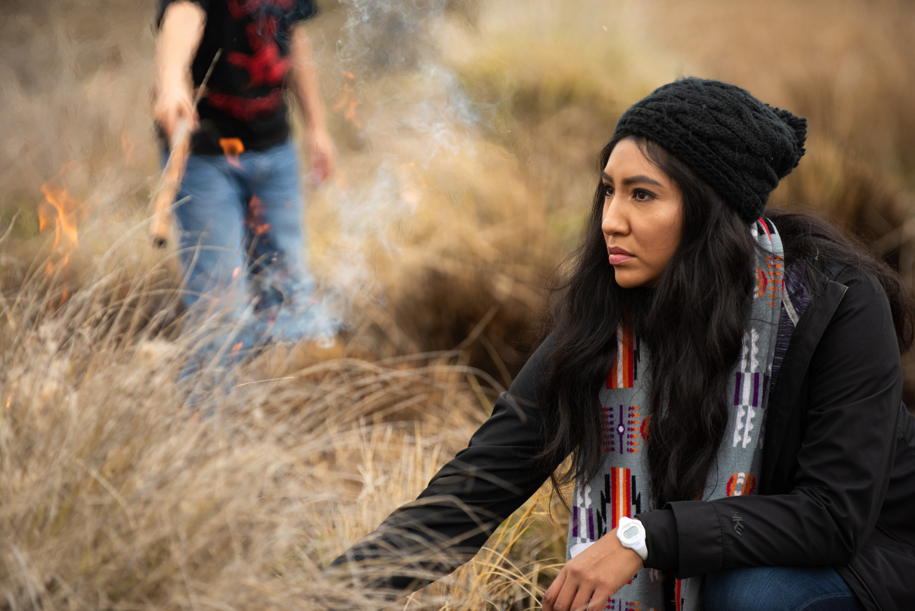 Melinda Adams wearing black beanie and black top squats down low to touch dry grasses in a field that is under going a control burn