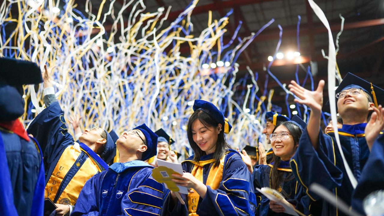 Graduate at Commencement looking down and smiling at program while others are looking up as confetti rains down.