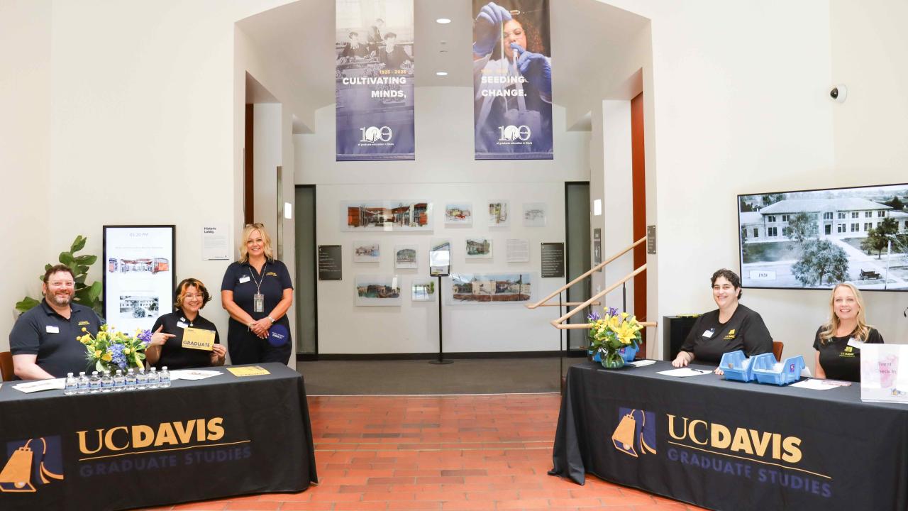 Smiling staff at two UC Davis Graduate Studies information tables in Walker Hall North Lobby with 100th Anniversary banners hanging above them.