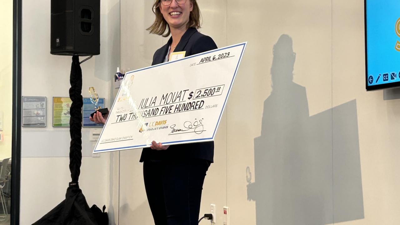Julia Mouat holds an oversized check in front of an LCD screen after winning 1st place in the UC Davis Grad Slam competition on April 6, 2023.