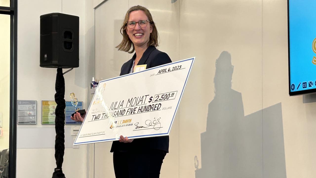Julia Mouat holds an oversized check in front of an LCD screen after winning 1st place in the UC Davis Grad Slam competition on April 6, 2023.