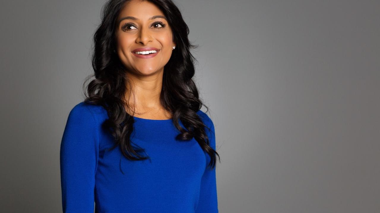Rinki Sethi, wearing a blue long sleeve blouse, looks off camera while smiling in front of a professional photography grey backdrop