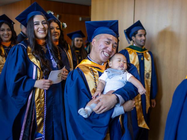 Graduate smiling with infant child at UC Davis Betty Irene Moore School of Nursing commencement.