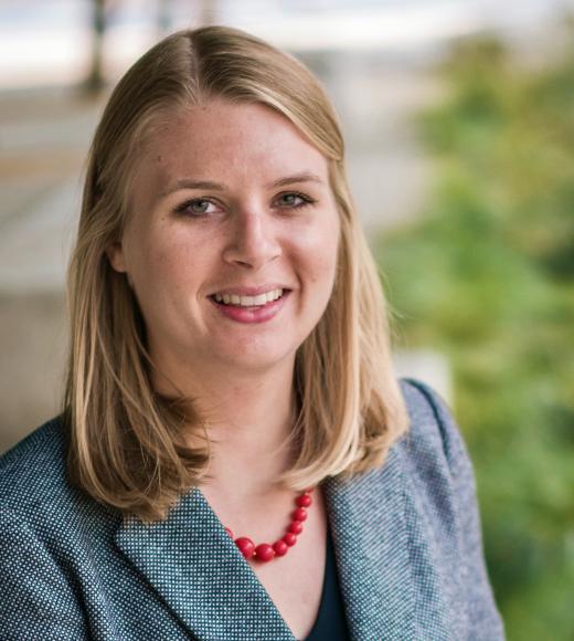 white woman with shoulder length blonde hair wears a grey sports coat and red beaded necklace.