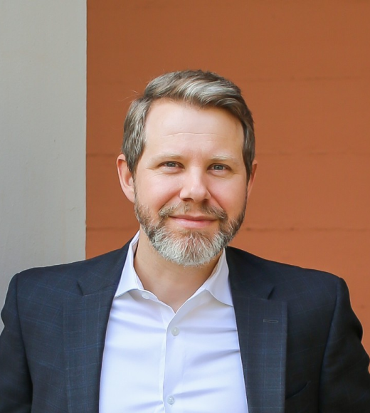 White man with greying hair and beard smiles at camera, wearing a white buttoned-down shirt and dark sports coat.light 