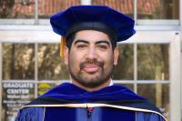 Smiling graduate in blue doctoral robes and velvet tam standing before the entrance to the Graduate Center at Walker Hall.