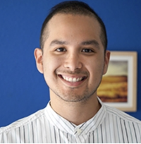 Jon Aguiñaga smiles at camera wearing a white shirt with a blue wall behind him.