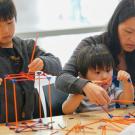 Two children build structures with kinex toys, their mother helps the youngest.