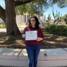 Dr. Jennifer Mogannam holds the Postdoctoral Excellence Award certificate outside the SCC with Rock Hall behind her on a sunny day