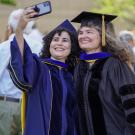 Graduate student in regalia smiling and taking selfie with mentor.