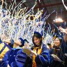Graduate at Commencement looking down and smiling at program while others are looking up as confetti rains down.