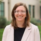 A woman with long brown hair and glasses smiles in front of a green background.