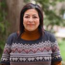 Woman with dark hair, wearing a patterned sweater, smiling outdoors in a green setting.