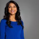 Rinki Sethi, wearing a blue long sleeve blouse, looks off camera while smiling in front of a professional photography grey backdrop